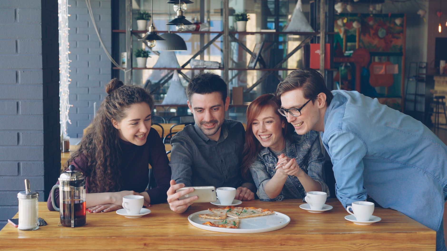 leaseradius-1786286051529 Four friends share a fun moment over coffee and pizza in a cozy cafe.
