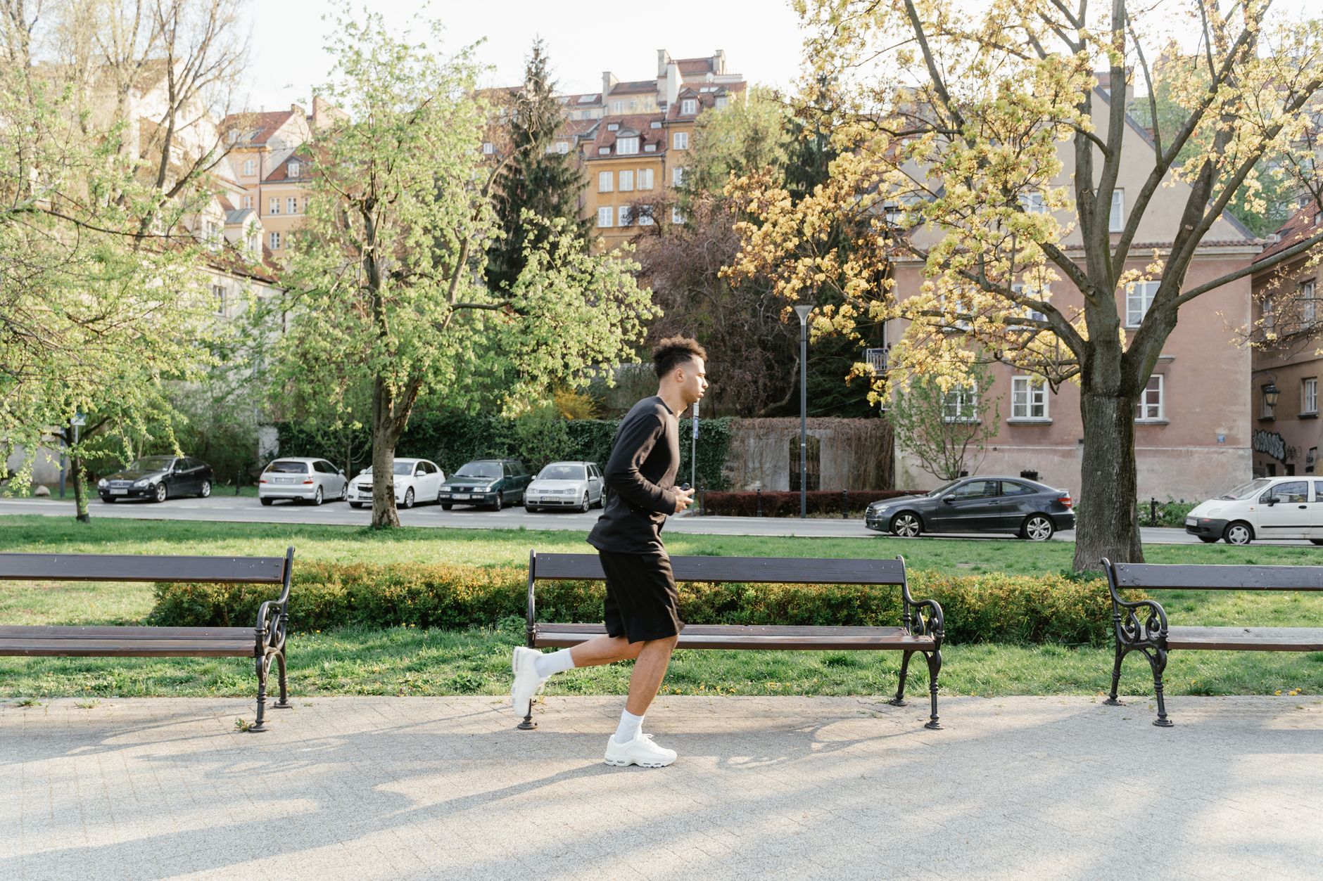 A man in athletic wear jogging through a sunlit park. Urban backdrop with trees and benches.