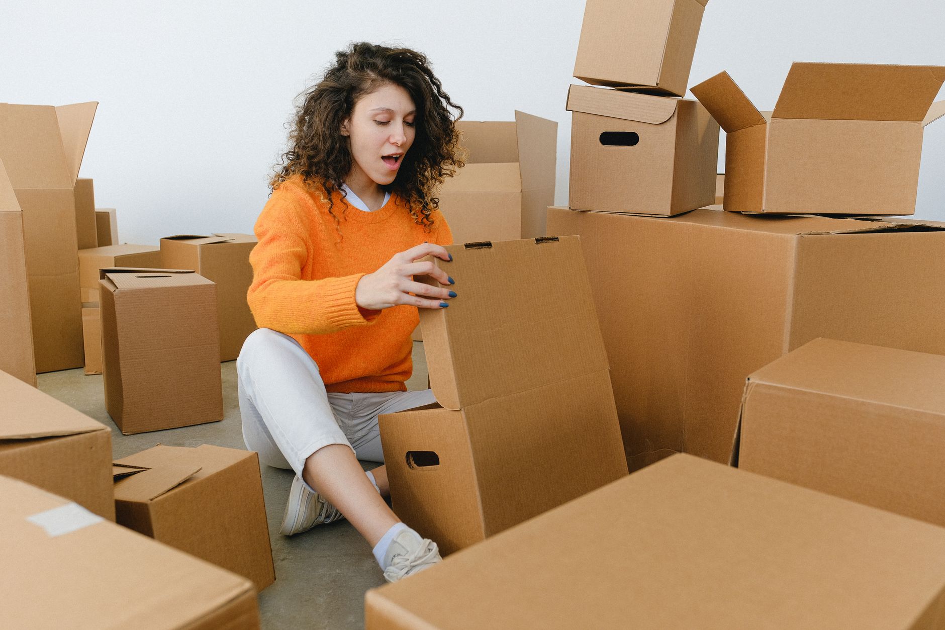 leaseradius-1788100447137 Amazed young female with curly hair in casual clothes sitting on floor and unpacking carton boxes with opened mouth after relocation
