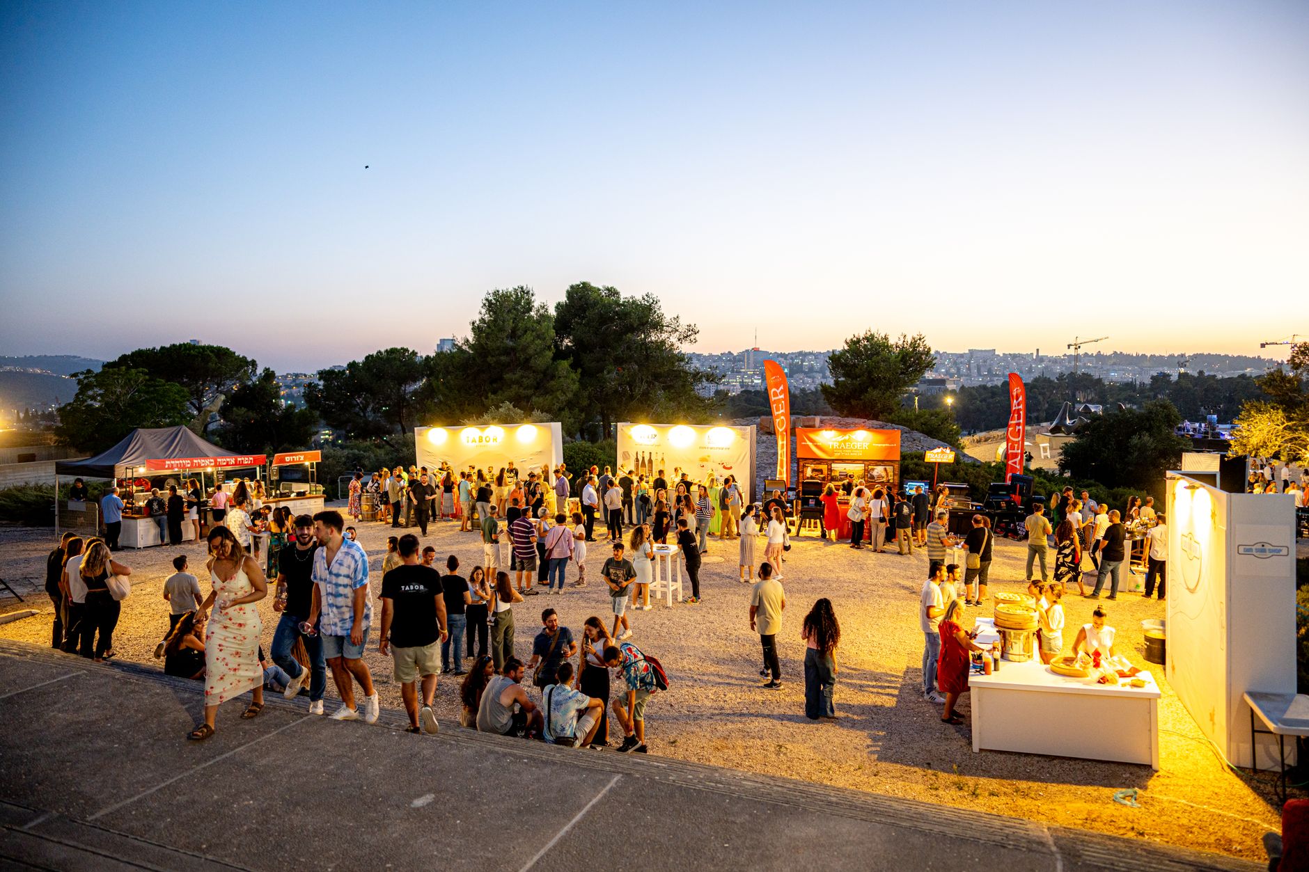 A lively outdoor festival at sunset with people socializing, food stalls, and a vibrant atmosphere.
