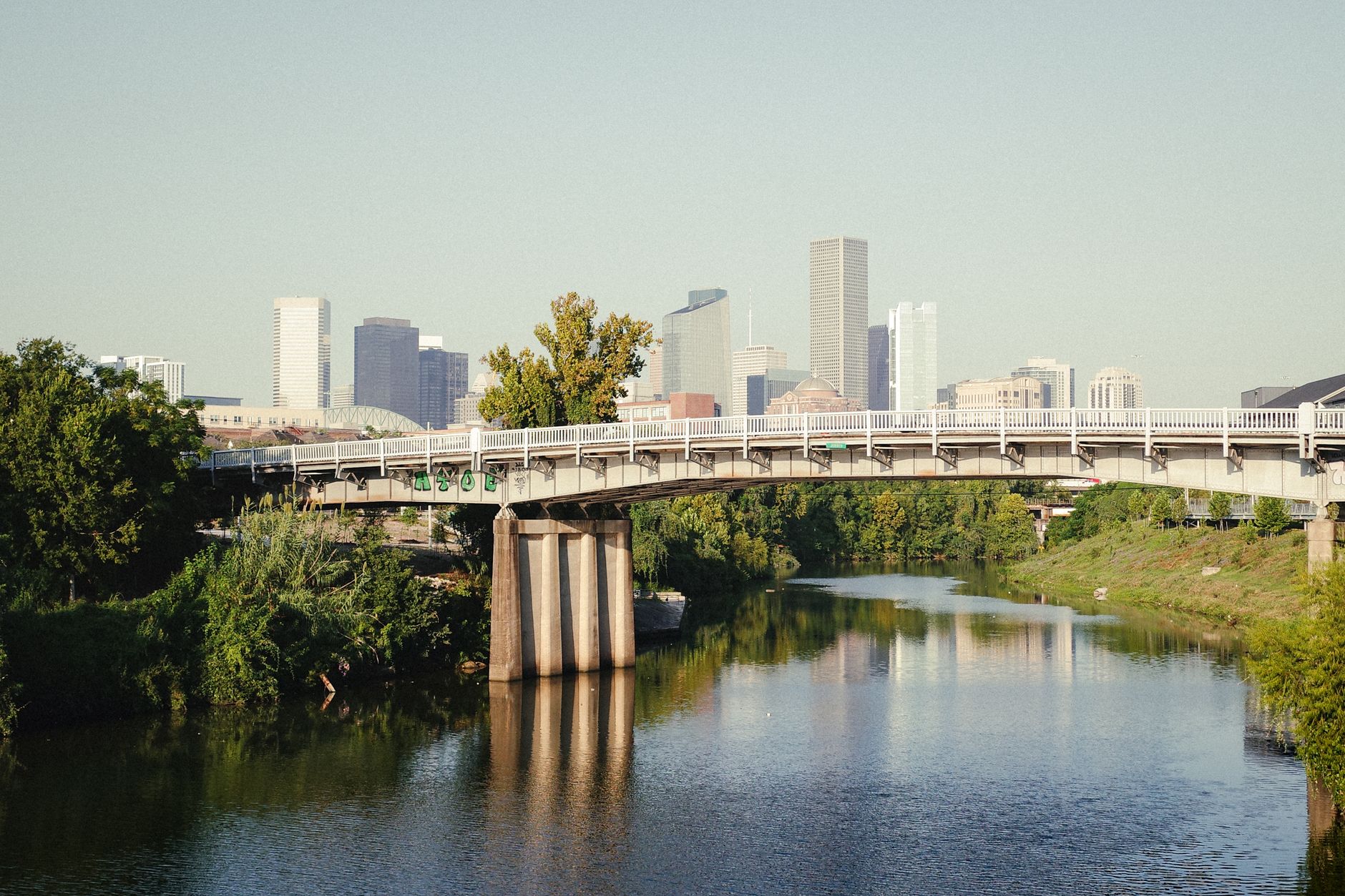 leaseradius-1791119598547 A tranquil view of Houston's skyline with a bridge over a serene bayou, surrounded by lush greenery.