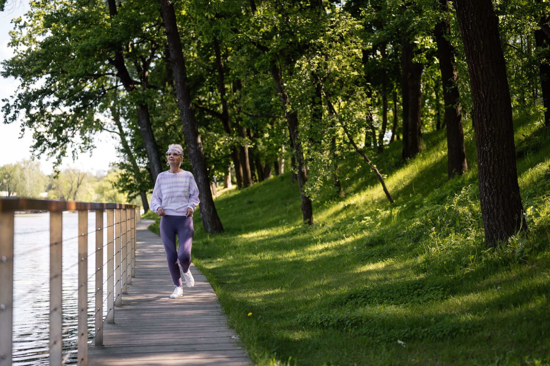 leaseradius-1792332127946 Elderly woman running along a riverside path in a green park, embracing fitness and nature.